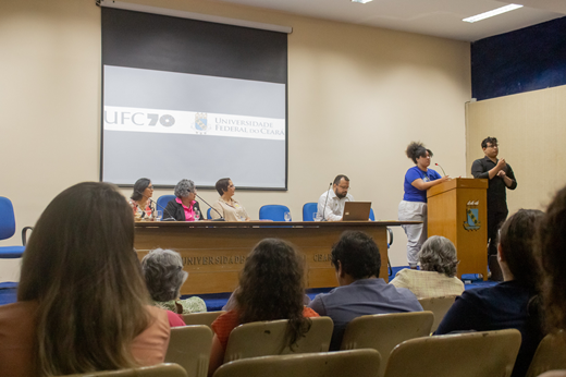 Imagem: No Auditório da Reitoria, Dandhara Cavalcante (estudante de História) discursa no púlpito para a plateia do evento Estatuinte em Debate; na mesa, estão Profª Irenísia Oliveira (presidente da ADUFC-Sindicato), Profª Diana Azevedo (vice-reitora), Elaine Teixeira (TAE) e Daniel Fonsêca (coord. de Articulação Política do Gabinete da Reitoria). 