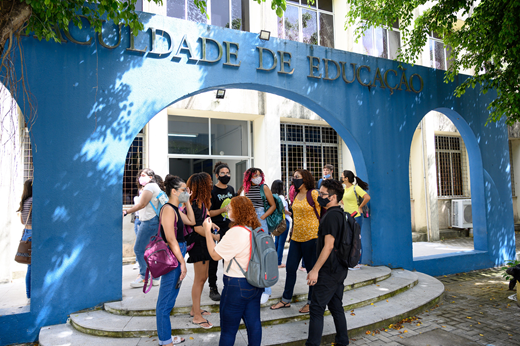 Imagem: Foto da fachada azul da faculdade de Educação mostra alunos conversdando na entrada.